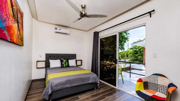 A modern bedroom with a bed, a ceiling fan, an air conditioning unit, and a view of a balcony with outdoor furniture.
