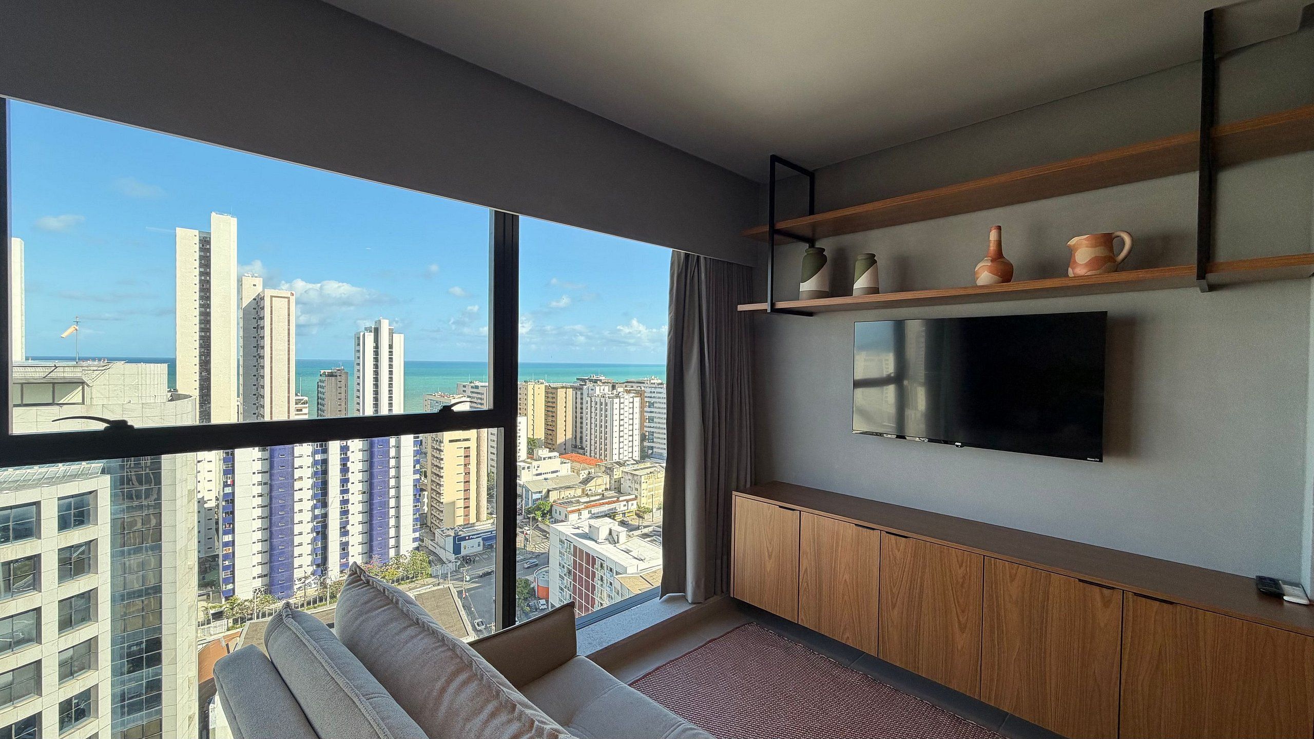 A modern apartment with a large balcony, a flat-screen TV mounted on the wall, and a view of the cityscape and ocean.