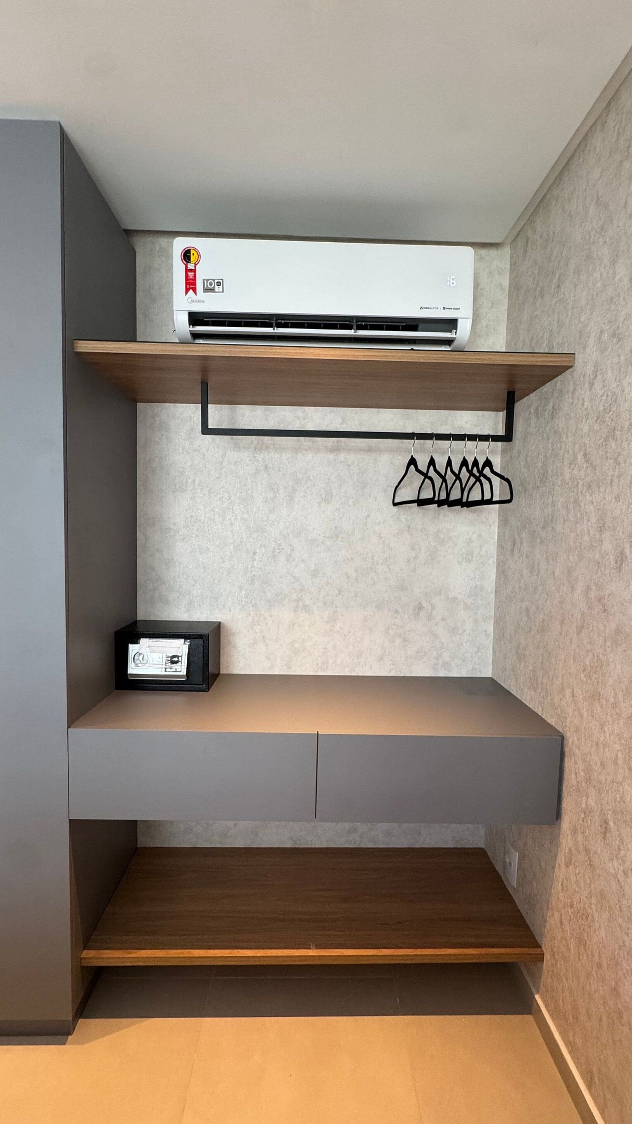 Una habitación con aire acondicionado integrado, un estante con perchero y un pequeño dispositivo electrónico en una repisa inferior.
