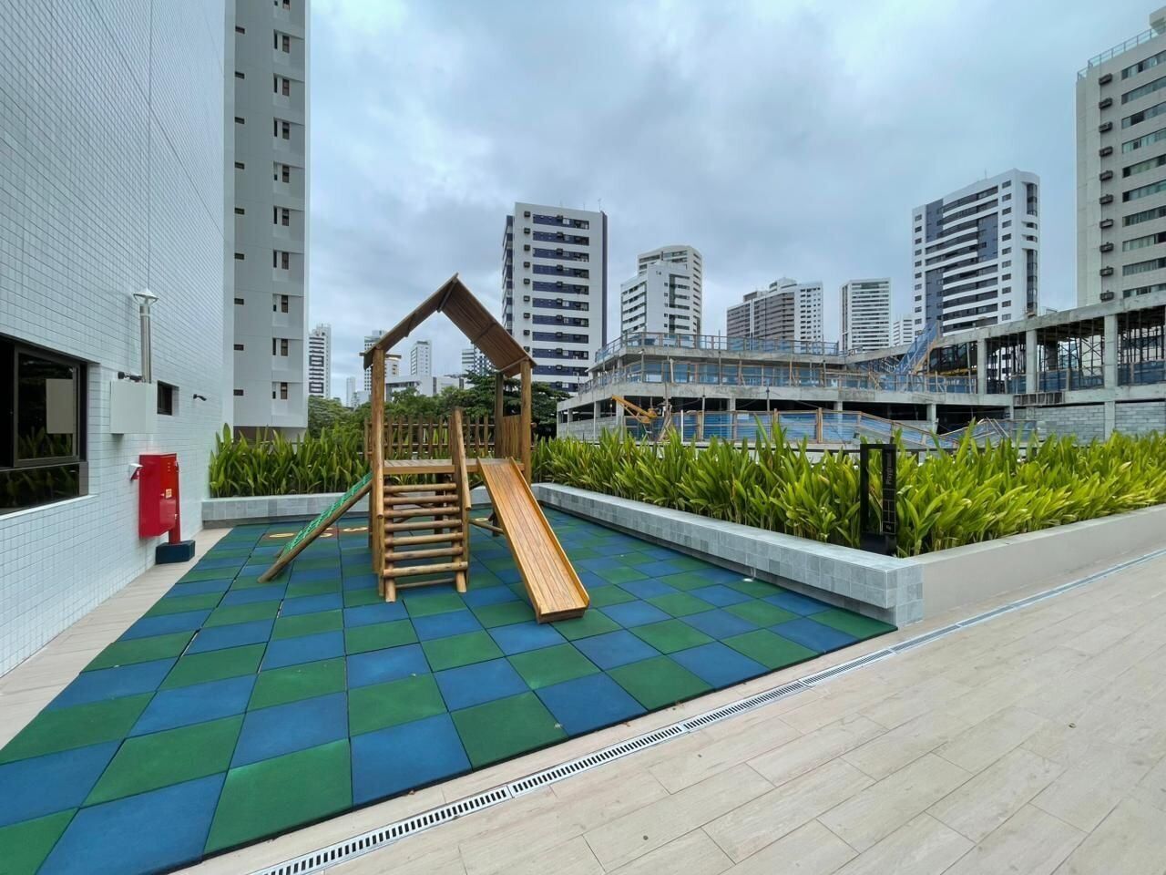 Zona de juegos al aire libre con un tobogán y una estructura de escalada, rodeada de un suelo de baldosas y un muro bajo con plantas.