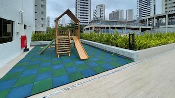 Zona de juegos al aire libre con un tobogán y una estructura de escalada, rodeada de un suelo de baldosas y un muro bajo con plantas.