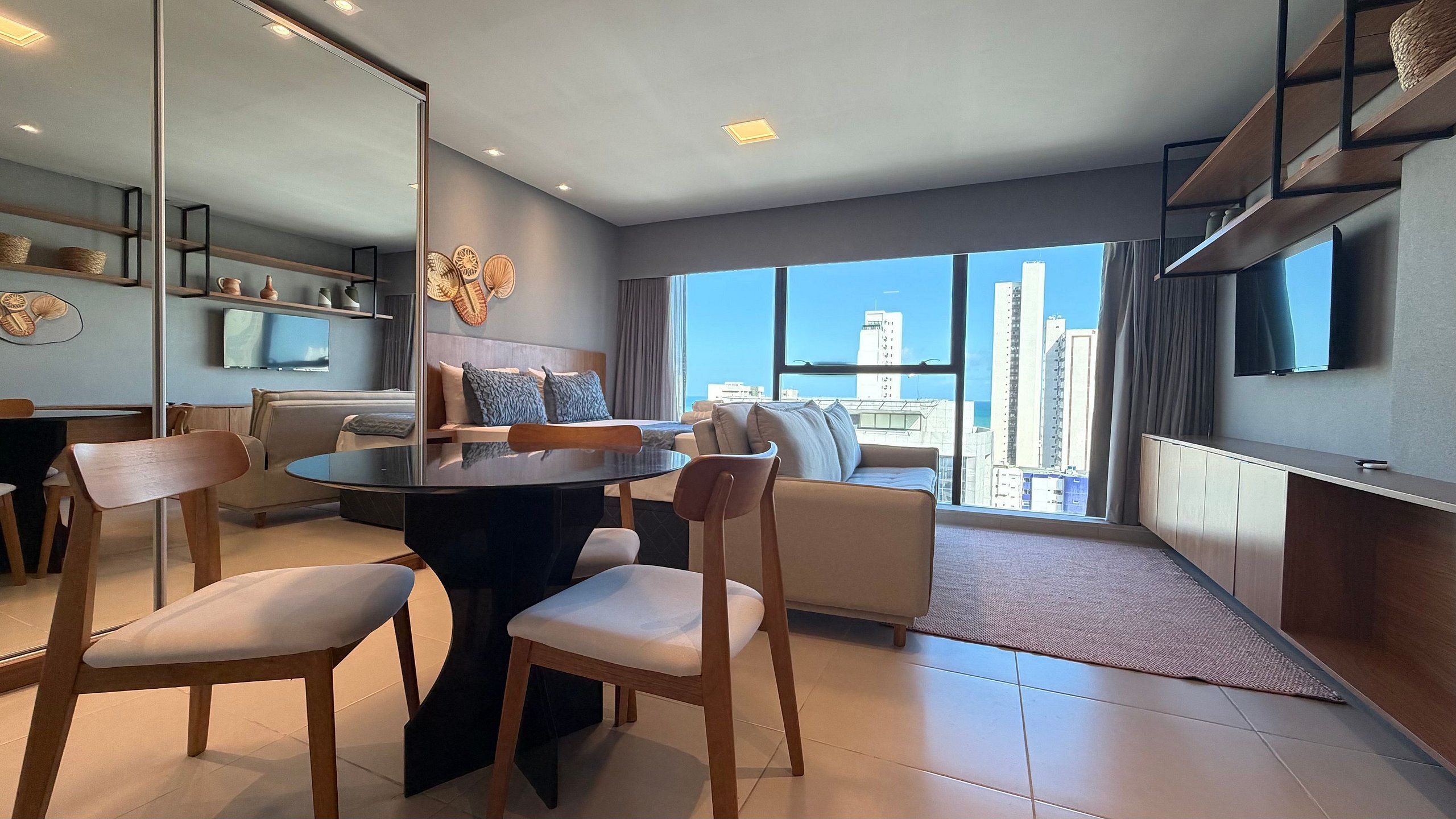 A modern hotel room with a dining area, a sofa, a TV, and a large window with a city view.