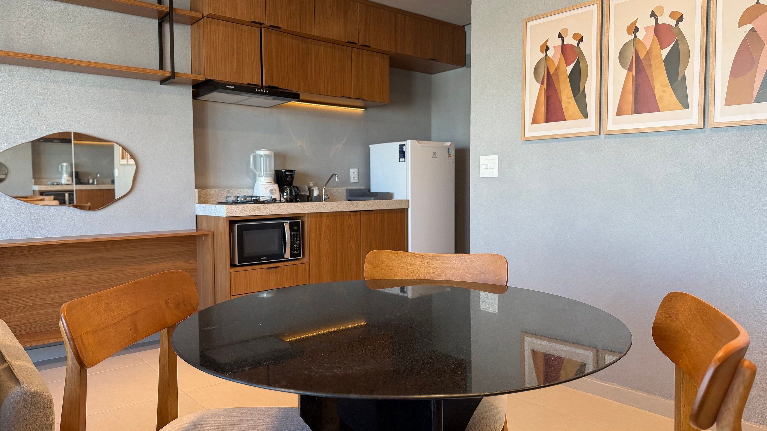 Una cocina moderna con armarios de madera, encimera negra, microondas, cafetera y refrigerador. Hay una mesa redonda con sillas y un espejo en la pared.