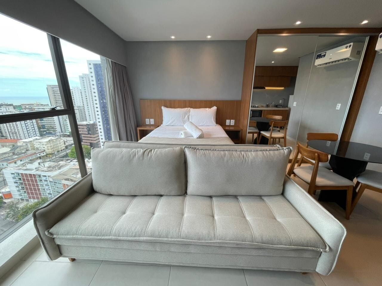 A modern hotel room with a sofa, a bed, a dining area, and a view of the city.