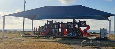 A playground with various play structures under a covered area, a picnic table, and a signboard.