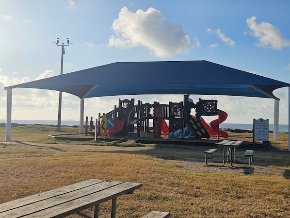 A playground with various play structures under a covered area, a picnic table, and a signboard.