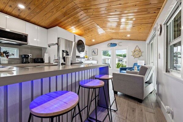 Une cuisine moderne avec un plafond en bois, un coin bar avec des tabourets et un espace salon avec un canapé et des fauteuils.