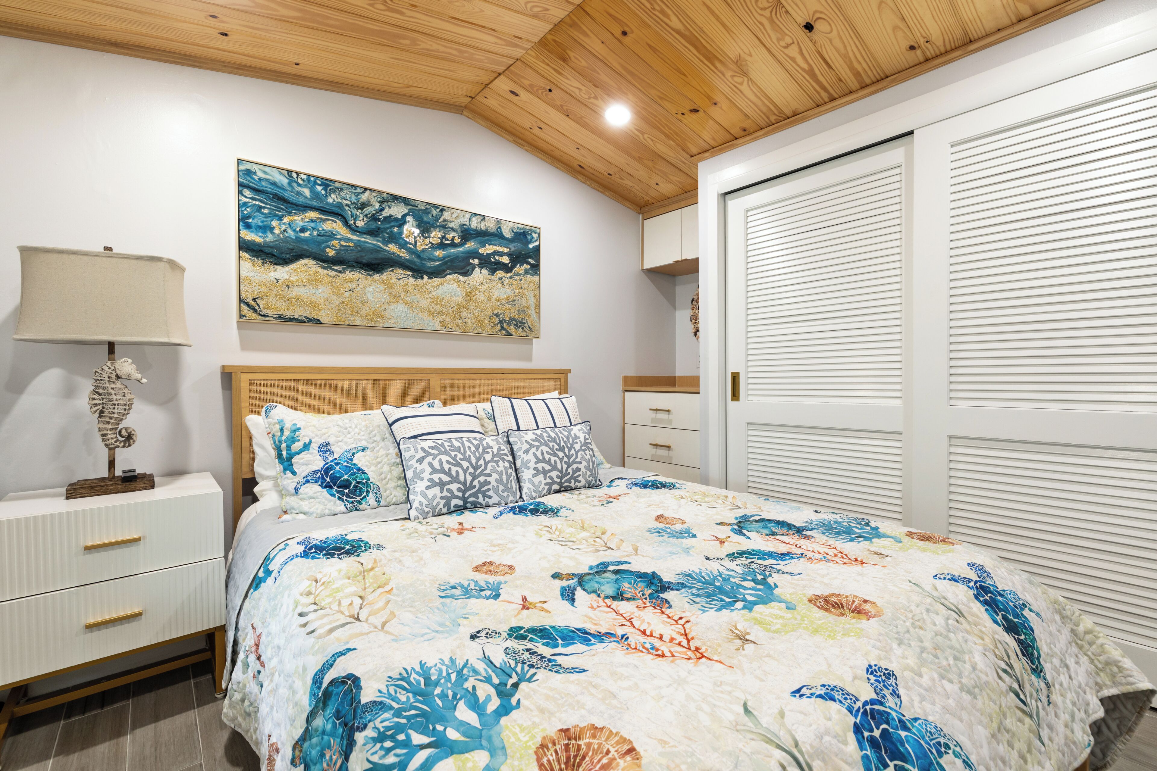 A bedroom with a wooden ceiling, a bed with a blue and white ocean-themed bedspread, a nightstand with a lamp, and a large painting above the bed.