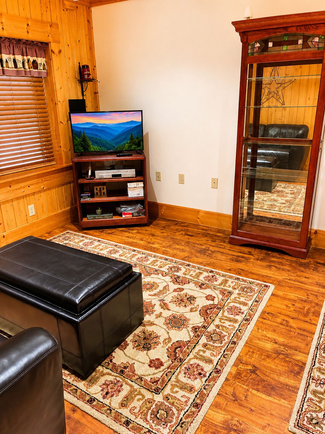 A cozy living room with a wooden floor, a television on a wooden stand, a leather ottoman, and a patterned rug.