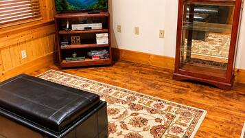 A cozy living room with a wooden floor, a television on a wooden stand, a leather ottoman, and a patterned rug.