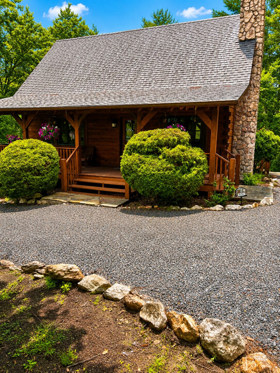 A wooden cabin with a stone chimney, a porch with steps, and two well-maintained bushes.