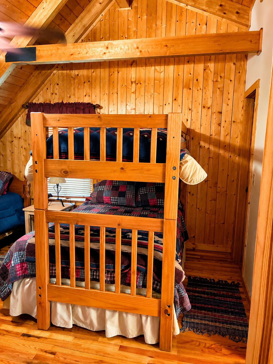 Una cama litera con estructura de madera, un escritorio con una lámpara y un sofá azul en una habitación con paredes y techo inclinados de madera.
