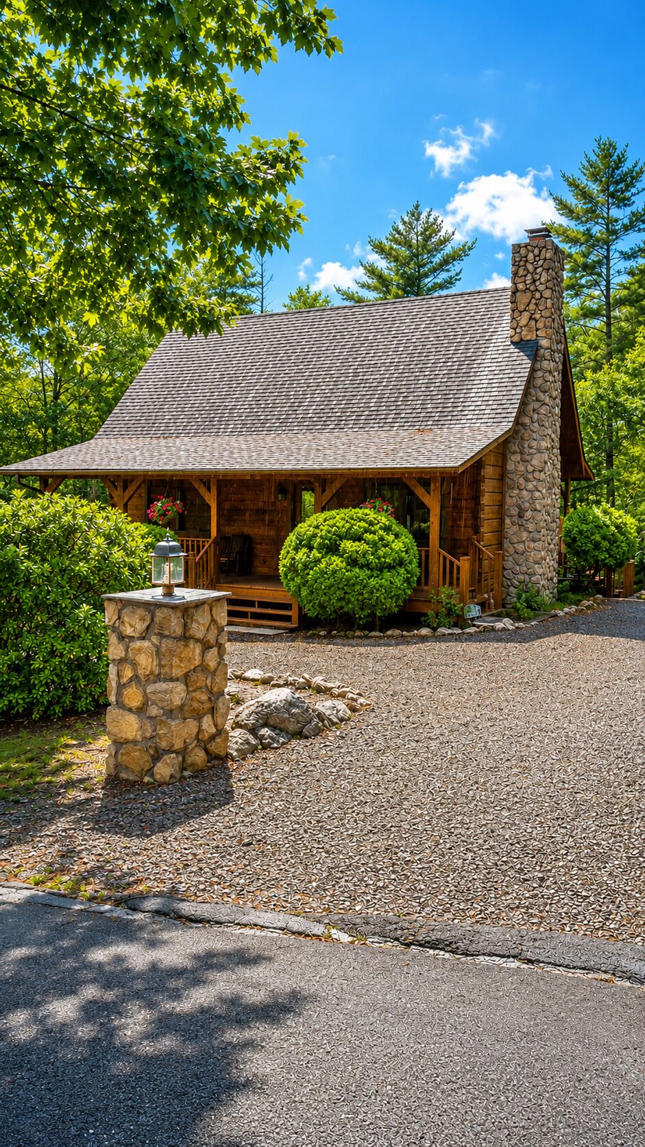 A log cabin with a stone chimney, surrounded by a gravel driveway and lush greenery.