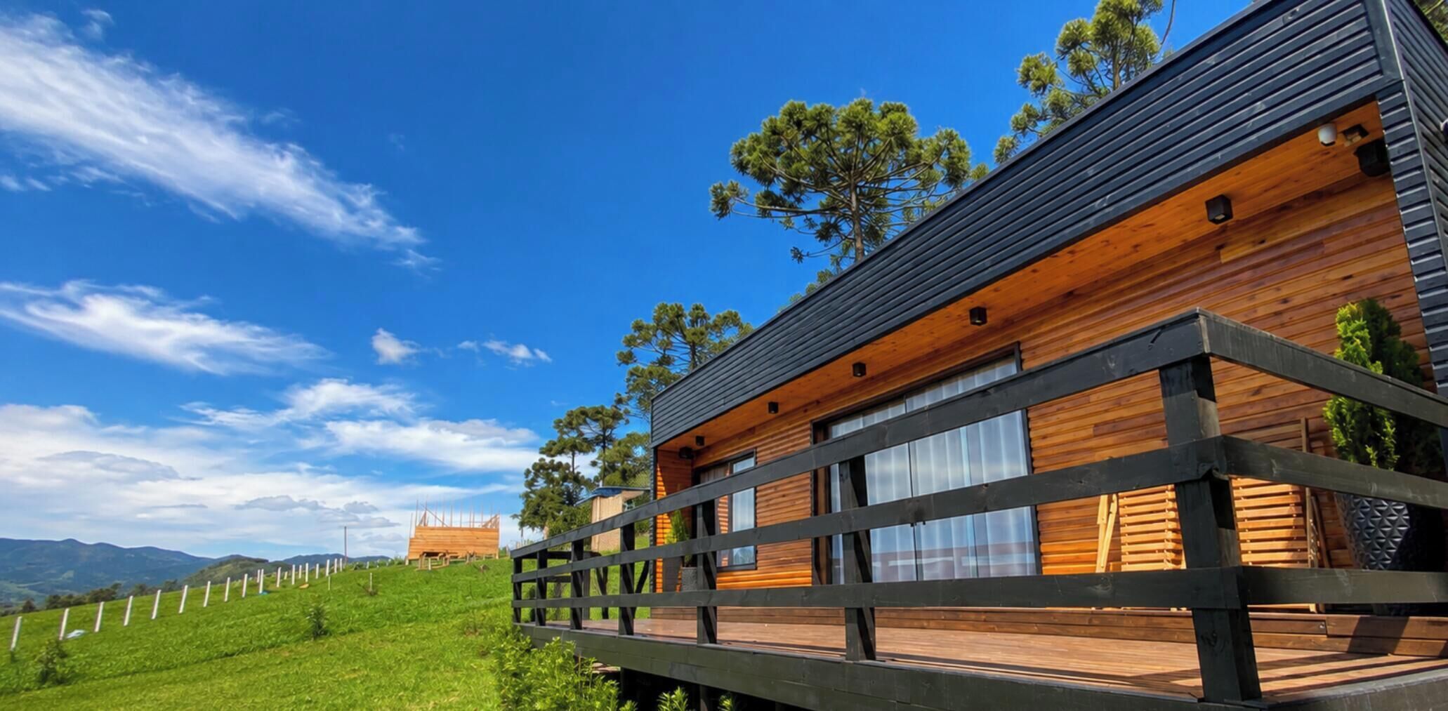 Une cabane en bois moderne, dotée d’une terrasse, entourée d’une clôture et d’arbres, se dressant sur un ciel dégagé.