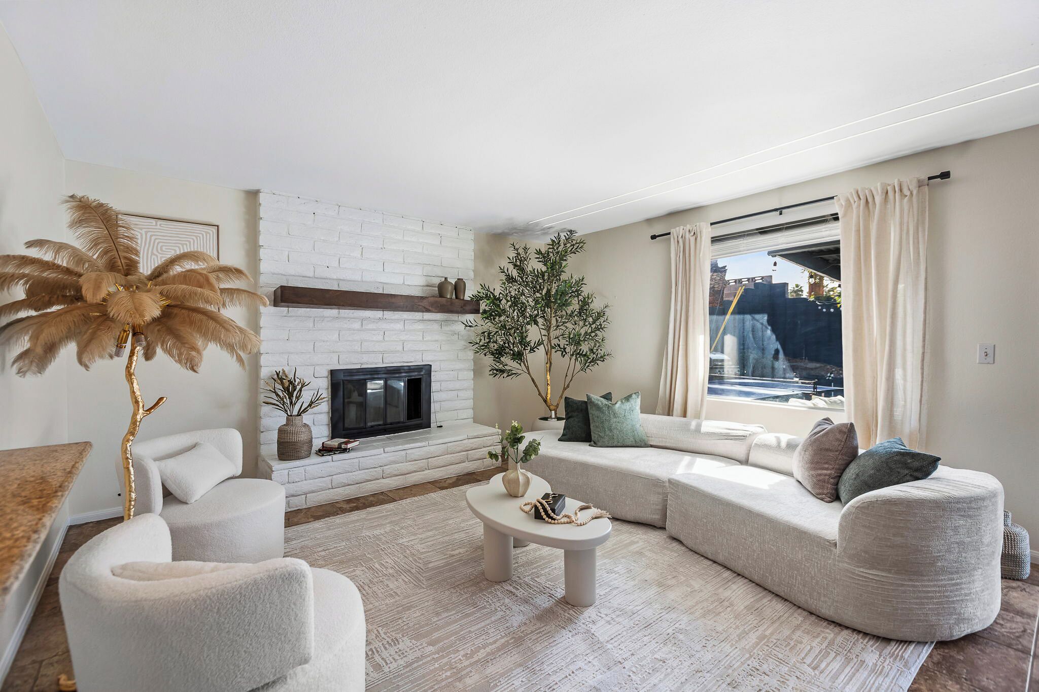 A modern living room with a fireplace, a large sofa, a coffee table, and a view through a window.