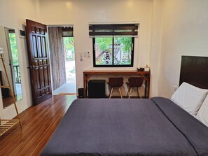 A bedroom with a bed, a desk with two chairs, a window with a view of greenery, and a wooden door. - Basilia Guest House (Santa Fe)