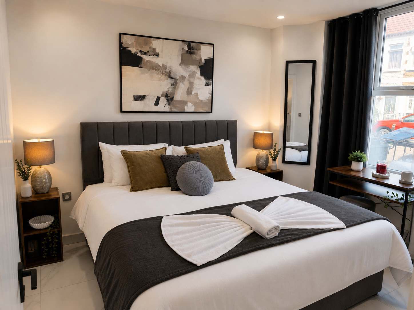 A hotel room with a large bed, two bedside tables with lamps, a wall-mounted abstract painting, a mirror, and a view of the street through the window.
