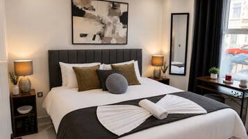 A hotel room with a large bed, two bedside tables with lamps, a wall-mounted abstract painting, a mirror, and a view of the street through the window.