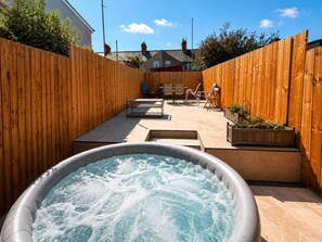 Ein Whirlpool in einem Garten mit Holzzaun, einer Terrasse mit Stühlen und Blick auf Häuser im Hintergrund.