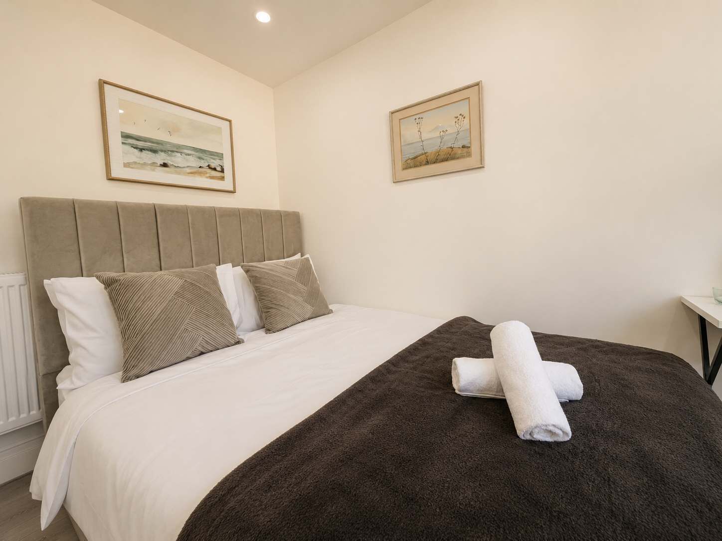 Een net opgemaakt bed met een bruine deken en twee kussens, een lijst met een landschapsfoto aan de muur, en een witte nachtkastje.