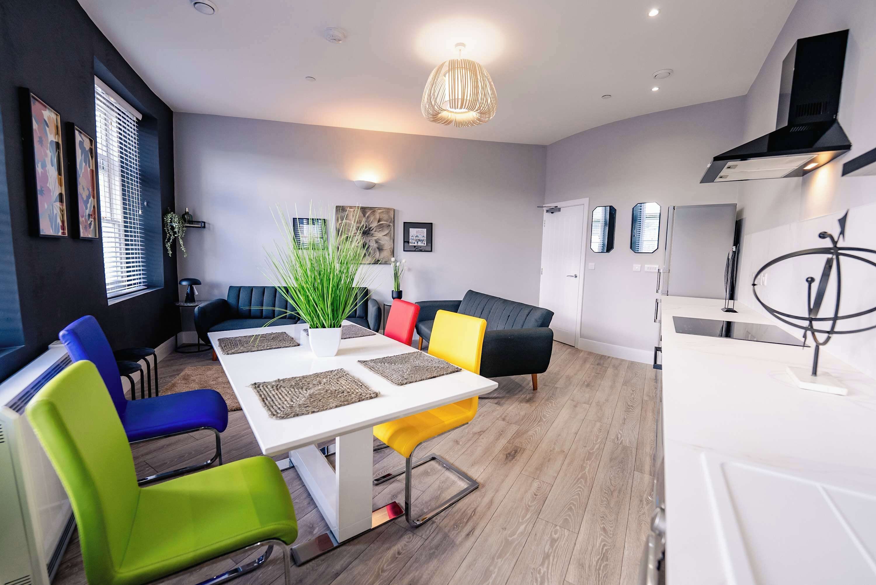 A modern living room with a dining table, colorful chairs, a sofa, and a kitchen area with a stove.
