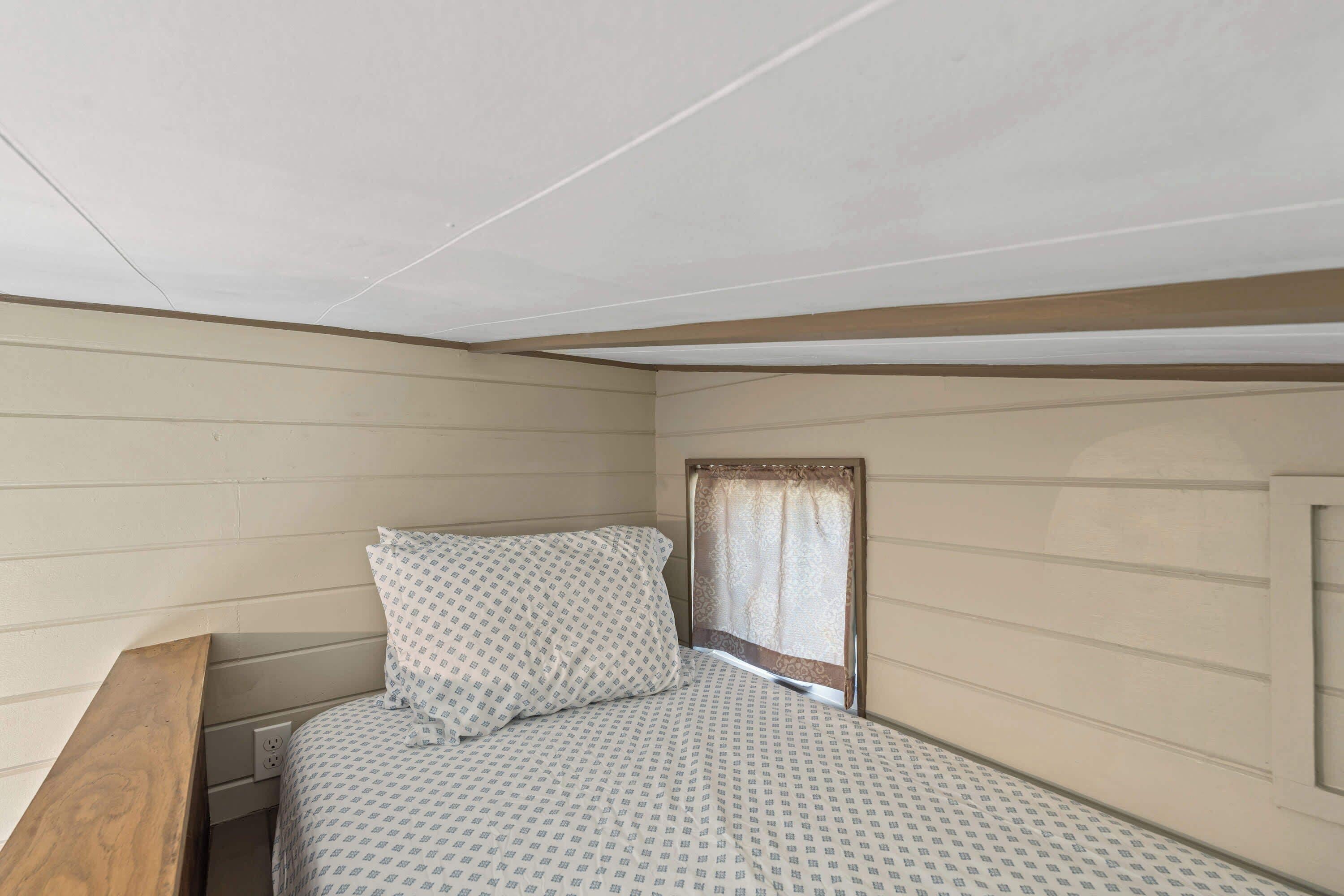 Une petite chambre avec un lit simple, une tête de lit en bois, une fenêtre avec un rideau et une prise électrique fixée au mur.
