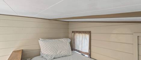 A small, single-bed room with a wooden headboard, a window with a curtain, and a wall-mounted electrical outlet.