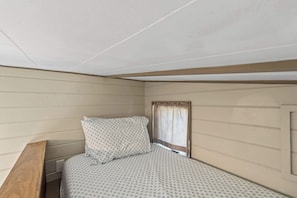 A small, single-bed room with a wooden headboard, a window with a curtain, and a wall-mounted electrical outlet. - NEW 2 bed tiny home on the farm (Asheville)