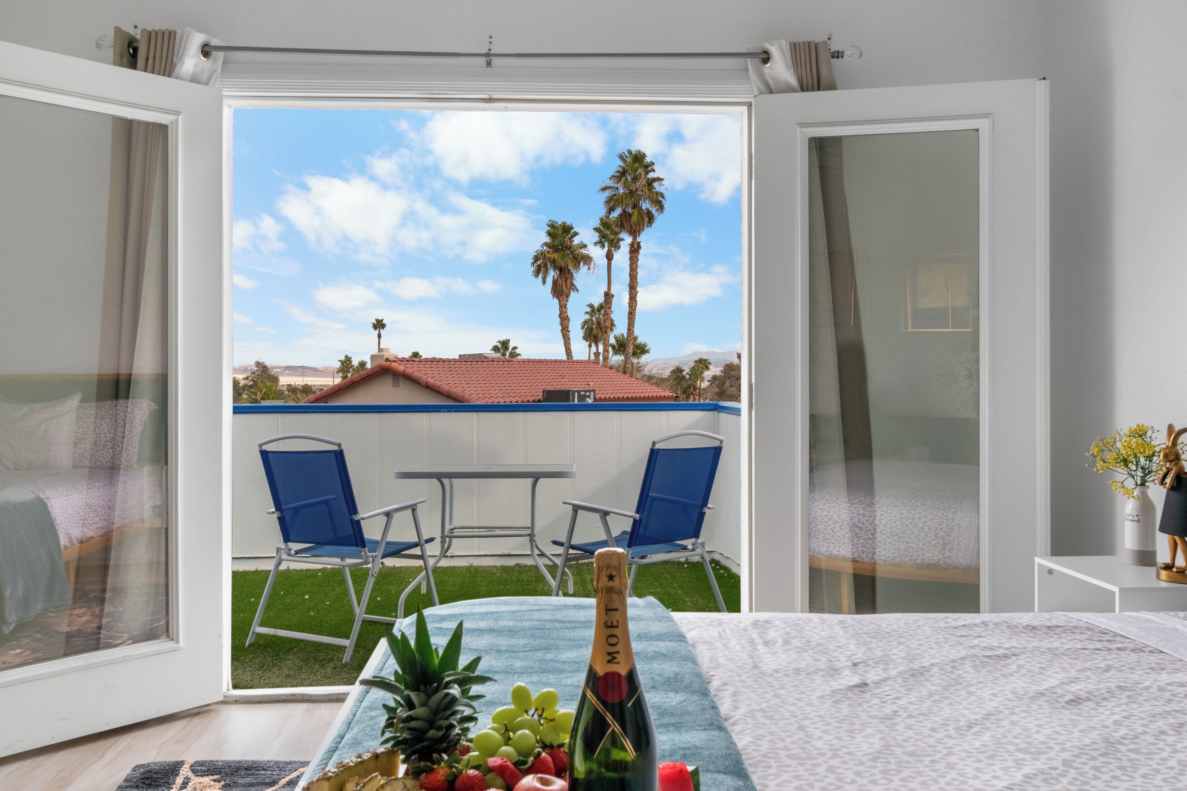 A room with a bed, a table with a bottle of champagne, and a view of a patio with chairs and palm trees.