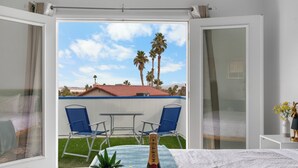 Ein Zimmer mit einem Bett, einem Tisch mit einer Champagnerflasche und Blick auf eine Terrasse mit Stühlen und Palmen.