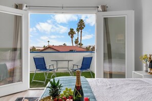 Ein Zimmer mit einem Bett, einem Tisch mit einer Champagnerflasche und Blick auf eine Terrasse mit Stühlen und Palmen.