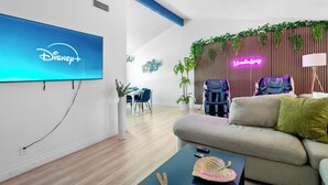 Ein modernes Wohnzimmer mit einem Flachbildfernseher, der das Disney+-Logo zeigt, einer Couch, einem Couchtisch mit einem dekorativen Gegenstand und einem Gaming-Sessel.