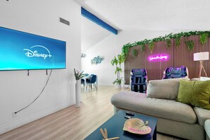 Ein modernes Wohnzimmer mit einem Flachbildfernseher, der das Disney+-Logo zeigt, einer Couch, einem Couchtisch mit einem dekorativen Gegenstand und einem Gaming-Sessel.