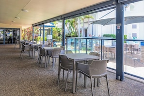 An outdoor seating area with tables and chairs, a glass railing, and a view of palm trees and buildings. - Mantra Top Floor Hotel Room (Urangan)