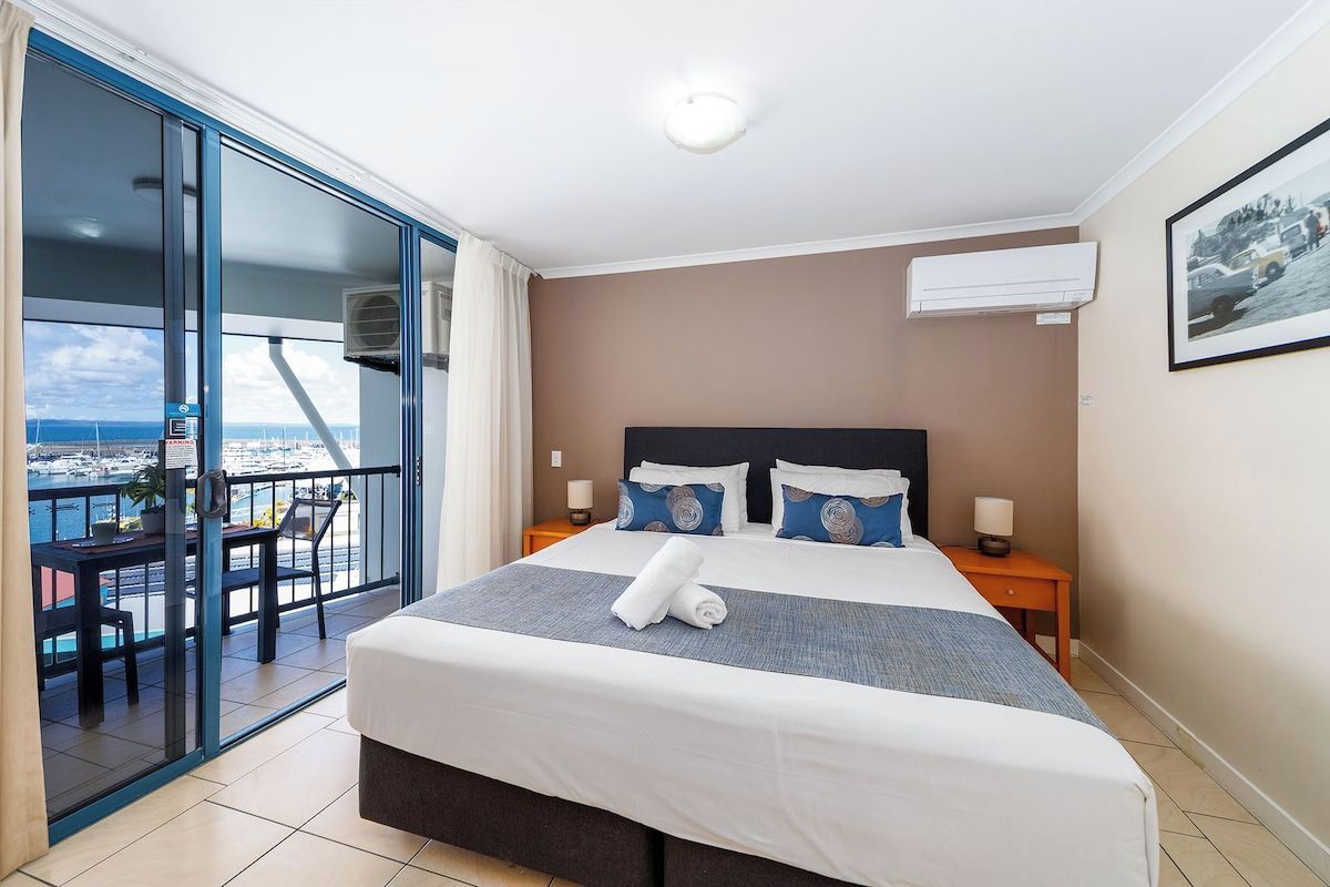 A hotel room with a bed, bedside tables, a balcony with a view, and an air conditioning unit.