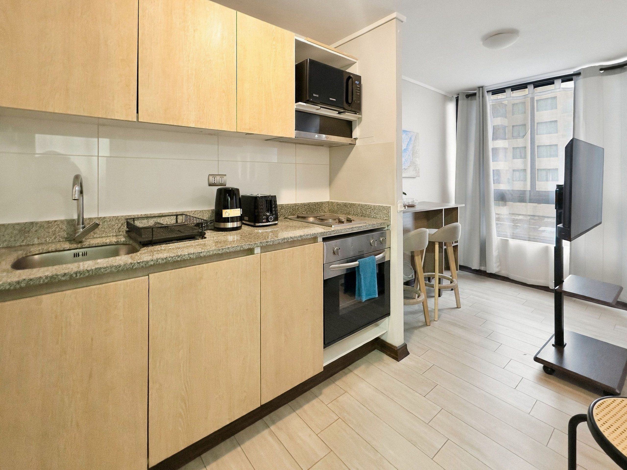 Una cocina moderna con microondas integrado, cafetera y horno. Hay un taburete y un mueble para televisión en la zona de estar contigua.