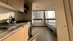 A compact kitchen with wooden cabinets, a microwave, and a coffee maker. There is a window with a view of buildings outside. - Oceana Suites Edificio Hipódromo  (Región Metropolitana)