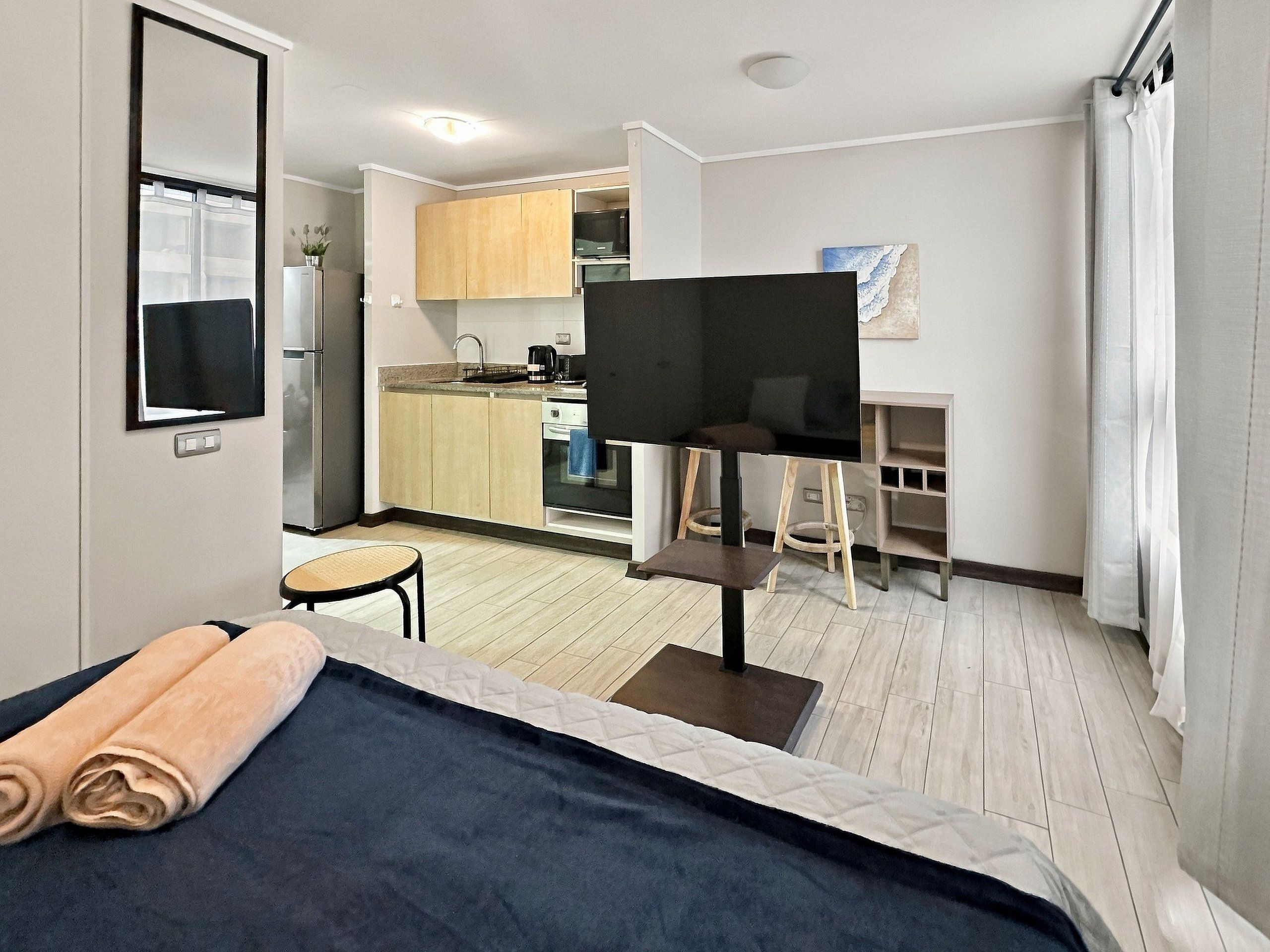 A modern hotel room with a bed, a small table, a TV, a kitchenette, and a bathroom.