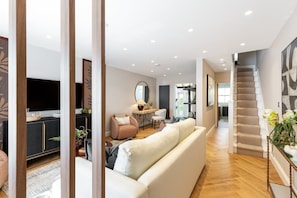 A modern living room with a sofa, a coffee table, and a television. There is a staircase leading to another level. - Luxury Three-Bed Chelsea Townhouse (London)
