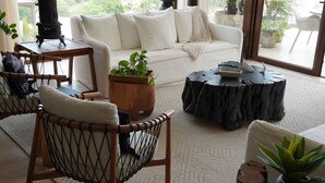 A cozy living room with a white sofa, a wooden coffee table, a round side table, and a wicker chair. There are potted plants and a large window providing a view of greenery outside. - Luxury penthouse with a private terrace and ocean views | Ayuuk (Cruz de Huanacaxtle)