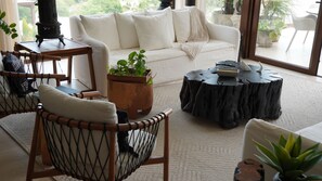 Ein gemütliches Wohnzimmer mit einem weißen Sofa, einem Holztisch, einem runden Beistelltisch und einem Korbstuhl. Zimmerpflanzen und ein großes Fenster bieten Blick auf Grünfläche draußen.