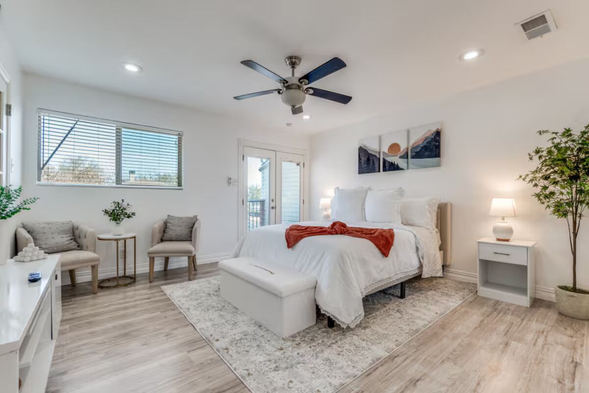 A bedroom with a bed, a ceiling fan, a bedside table, a nightstand, a window with blinds, and a potted plant.