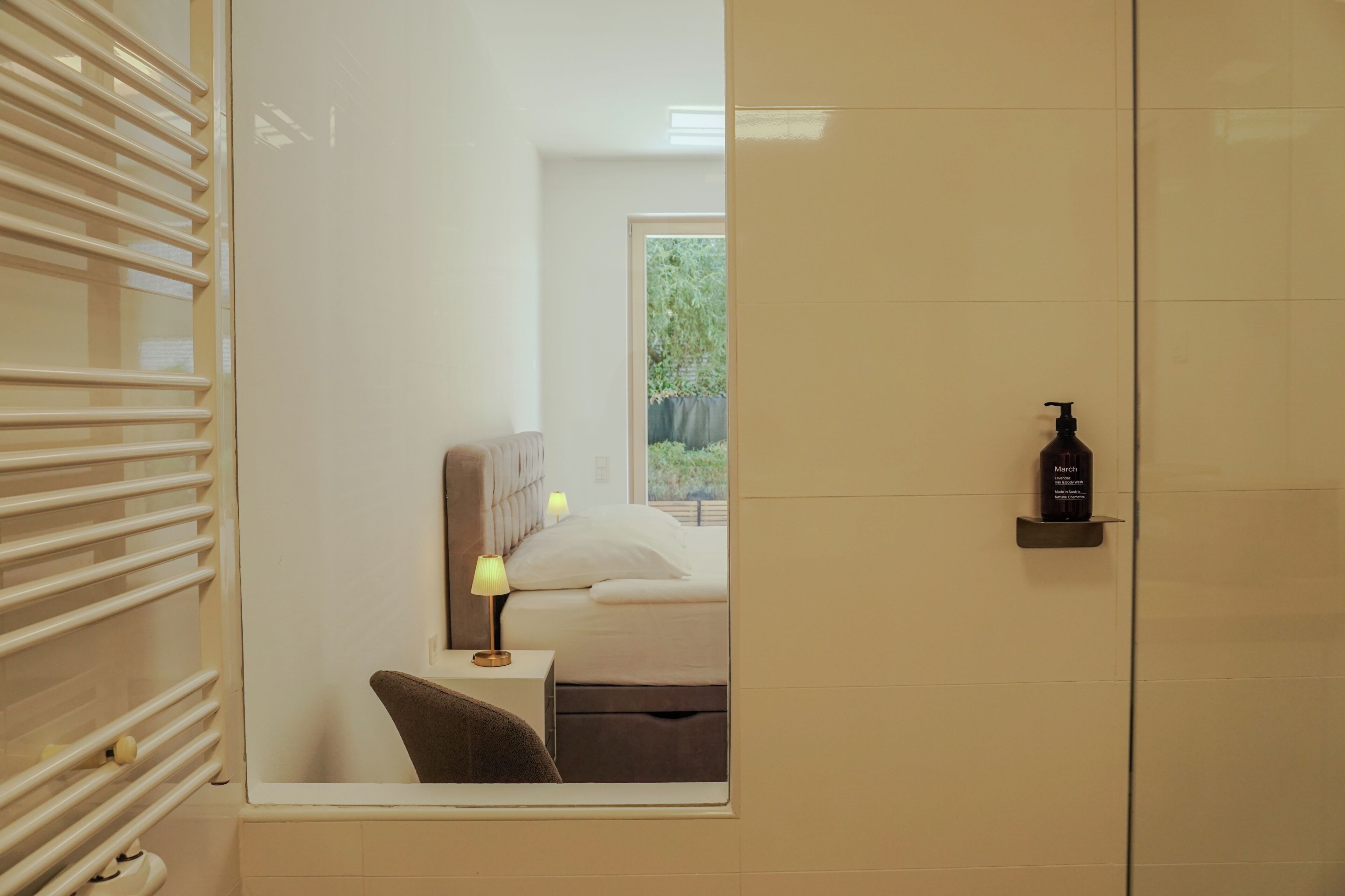 A bedroom with a bed, a nightstand, a lamp, and a bottle of March body wash on a shelf.