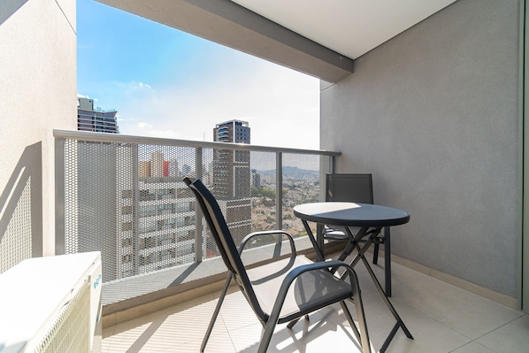 A balcony with a table and chairs, a mesh safety barrier, and a cityscape view.