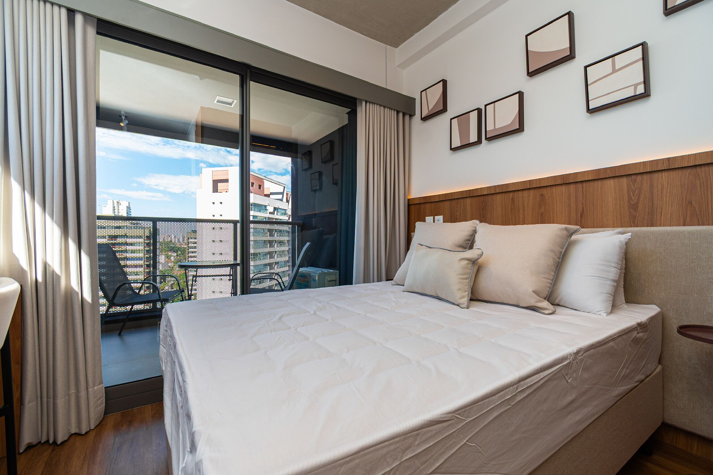 Una habitación de hotel con una cama grande, un cabecero de madera, un balcón con vistas a edificios y varias fotografías enmarcadas en la pared.