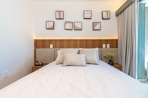 A hotel room with a large bed, a wooden headboard, a nightstand with a bottle and a candle, and a window with curtains.