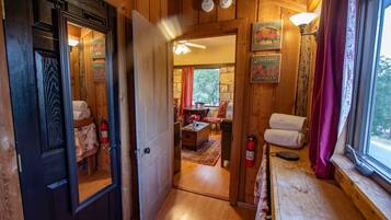 A wooden interior with a door leading to a room with a bed, a table, and a chair. There are wall-mounted light fixtures and a window with curtains.