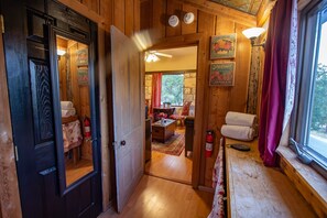 Een houten interieur met een deur die leidt naar een kamer met een bed, een tafel en een stoel. Er zijn wandlampen en een raam met gordijnen.