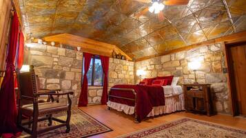 A bedroom with a stone wall, a wooden ceiling, a bed with red bedding, a chair, and a nightstand.