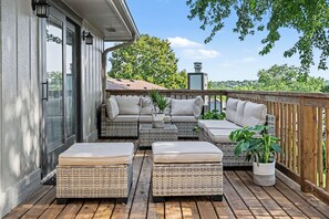 A covered outdoor seating area with wicker furniture and cushions, a wooden deck, and a view of the surrounding landscape.